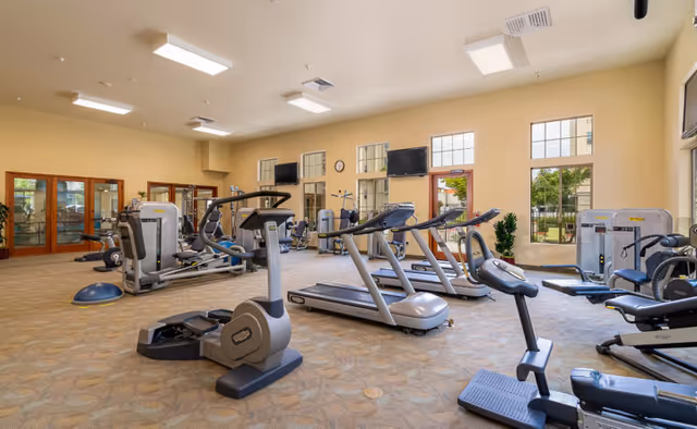 Bright fitness room with treadmills, stationary bikes, and weight machines in a senior living facility.
