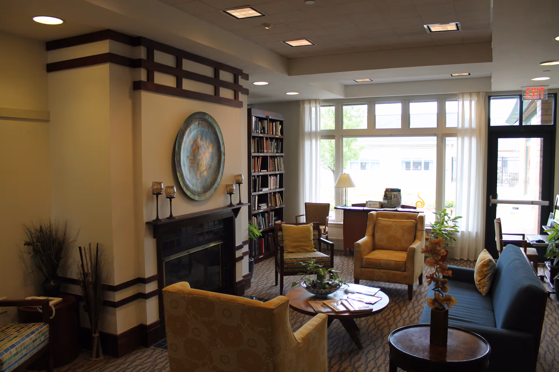 A cozy living room area in The Summit at Brighton Independent Living featuring a fireplace with decorative candles and a round wall art above it. The room has comfortable seating including two yellow armchairs, a dark blue sofa, and a wooden chair. A round wooden coffee table with plants and magazines is in the center. There is a bookshelf filled with books, a desk with a lamp near large windows with white curtains, and an exit door to the right.