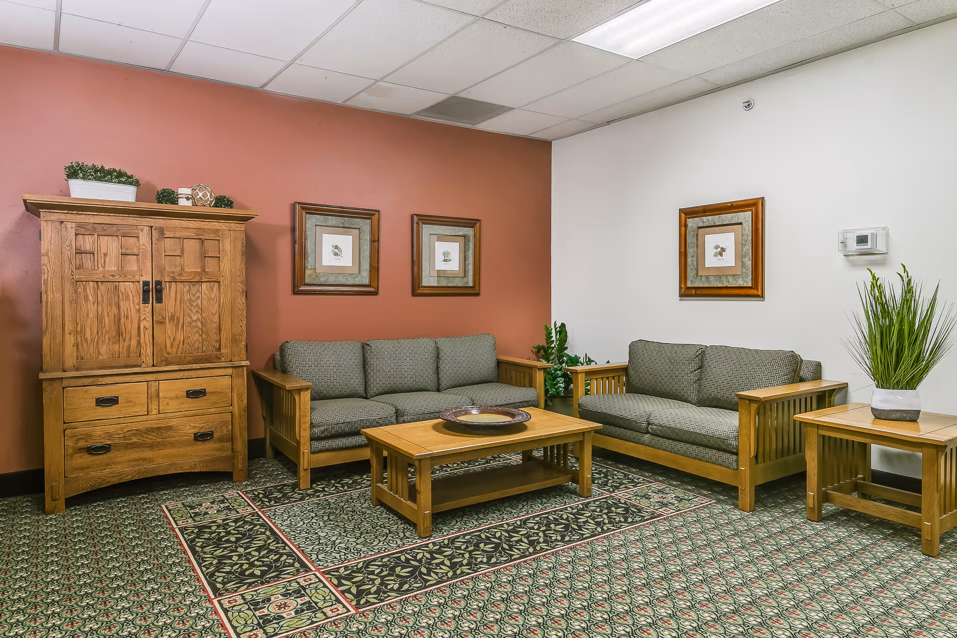A cozy living room area with two wooden framed sofas with gray cushions, a wooden coffee table with a decorative plate, a wooden side table with a potted plant, and a wooden cabinet with plants on top. The walls are painted white and a warm reddish-brown color, adorned with framed botanical prints. The floor is covered with a patterned carpet.