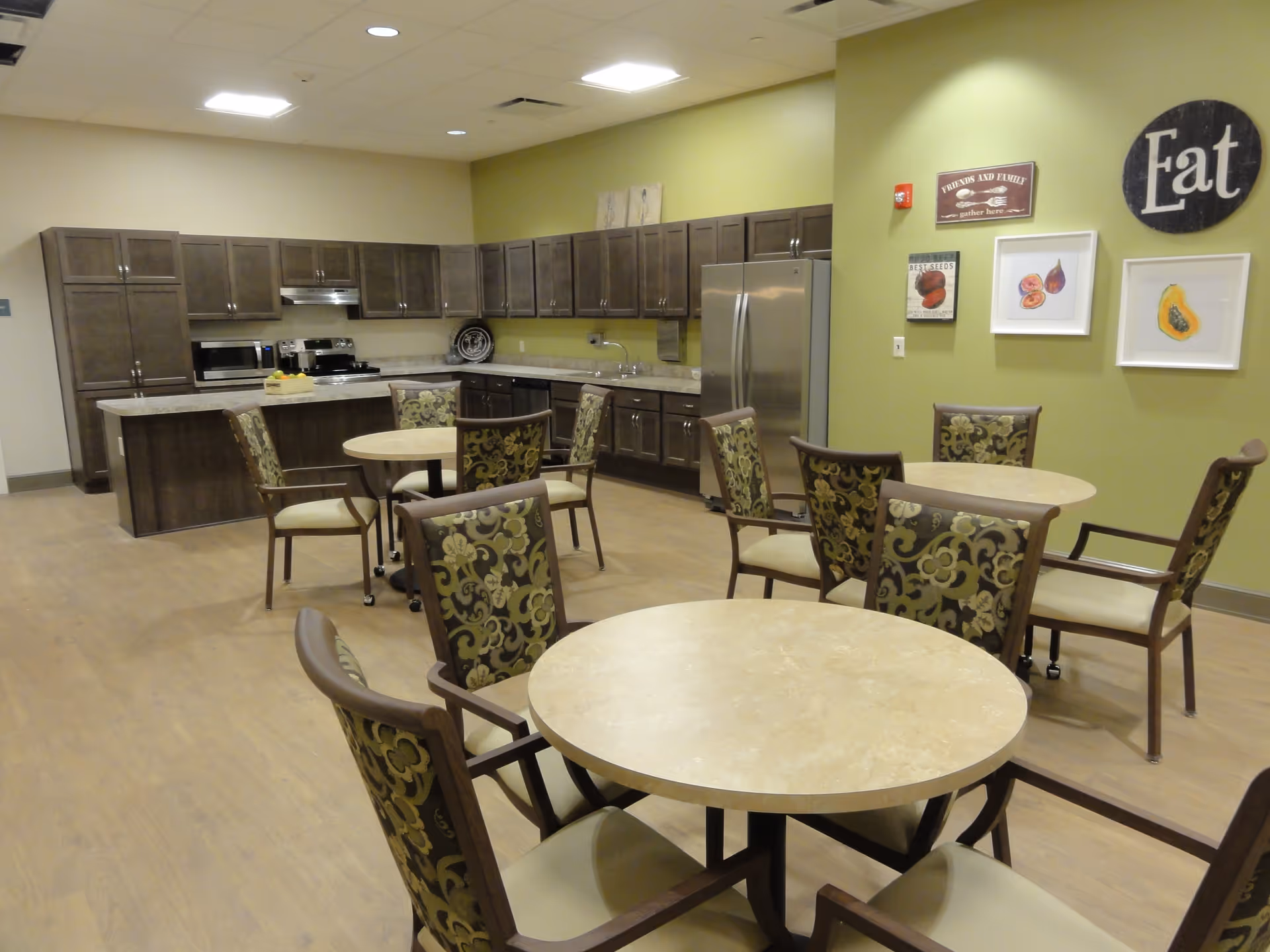 A communal kitchen and dining area with round tables and patterned chairs. The kitchen has dark wood cabinets, a stainless steel refrigerator, microwave, stove, and a countertop with a fruit basket. The walls are painted green and decorated with framed pictures of fruits and a round sign that says 'Eat'.