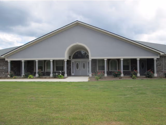 Front exterior view of a single-story building with a gray roof and light gray facade, featuring a covered entrance with white columns and double doors, surrounded by a well-maintained lawn and some small bushes.