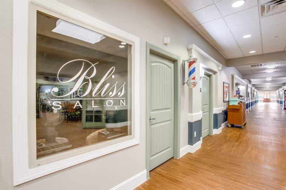 Interior hallway of The Landing at Stone Oak Memory Care with light green doors and wood flooring. A large window on the left displays the words 'Bliss Salon'. A traditional barber pole is mounted on the wall next to one of the doors. The hallway is well-lit with ceiling lights and has a cart with supplies further down the corridor.