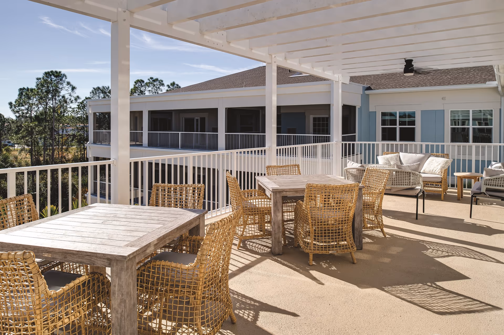 Outdoor covered patio area with wooden tables and wicker chairs, adjacent to a building with blue and white exterior walls and windows. The patio has a white pergola roof and overlooks some trees and greenery.