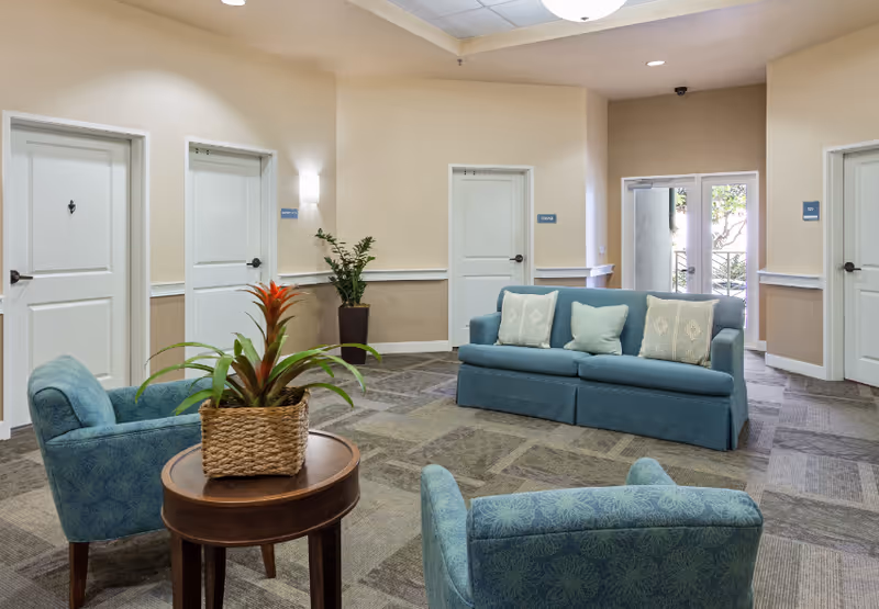 A seating area in a senior living facility with a blue sofa and two blue armchairs arranged around a small round wooden table with a potted plant. The room has beige walls, carpeted floor, several white doors, and a glass door leading outside.