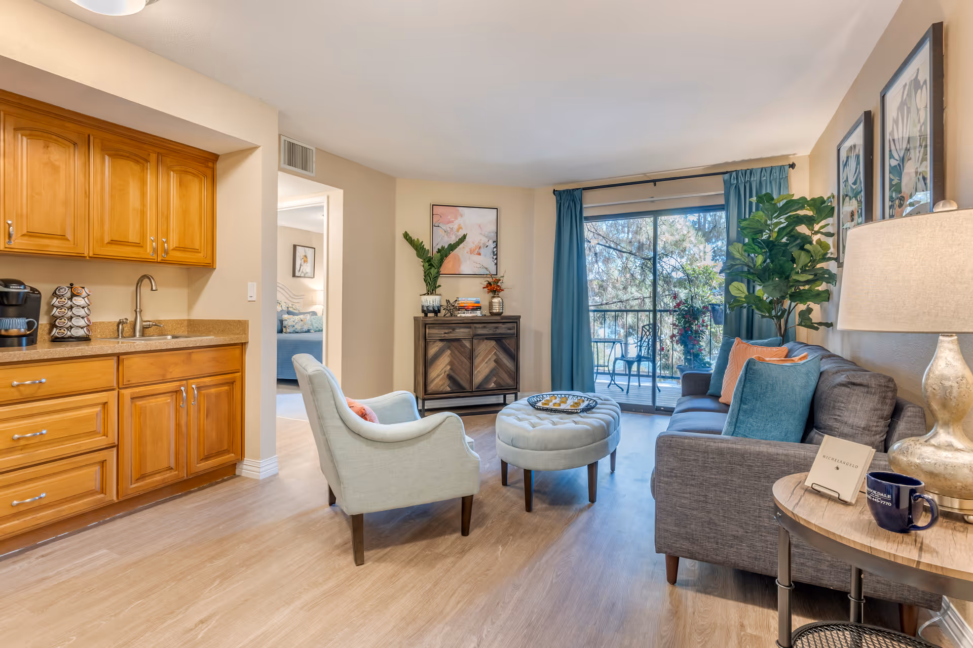 A cozy living room area in a senior living facility with a gray sofa adorned with colorful pillows, a light blue armchair, and a round tufted ottoman in the center. There is a wooden cabinet against the wall with decorative items and a framed artwork above it. Large sliding glass doors with blue curtains open to a balcony with outdoor seating and greenery visible outside. To the left, there is a kitchenette with wooden cabinets, a countertop, a sink, and a coffee maker.