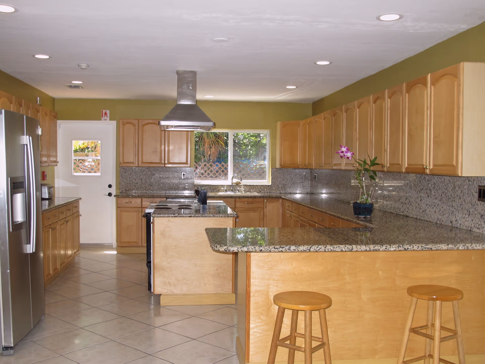 A spacious kitchen with light wood cabinets and granite countertops. The kitchen features a large island with a stovetop and a stainless steel range hood above it. There is a stainless steel refrigerator on the left, a window above the sink at the back, and two wooden stools at the counter on the right. The floor is tiled with large square tiles, and the walls are painted green. A small potted orchid is placed on the counter.