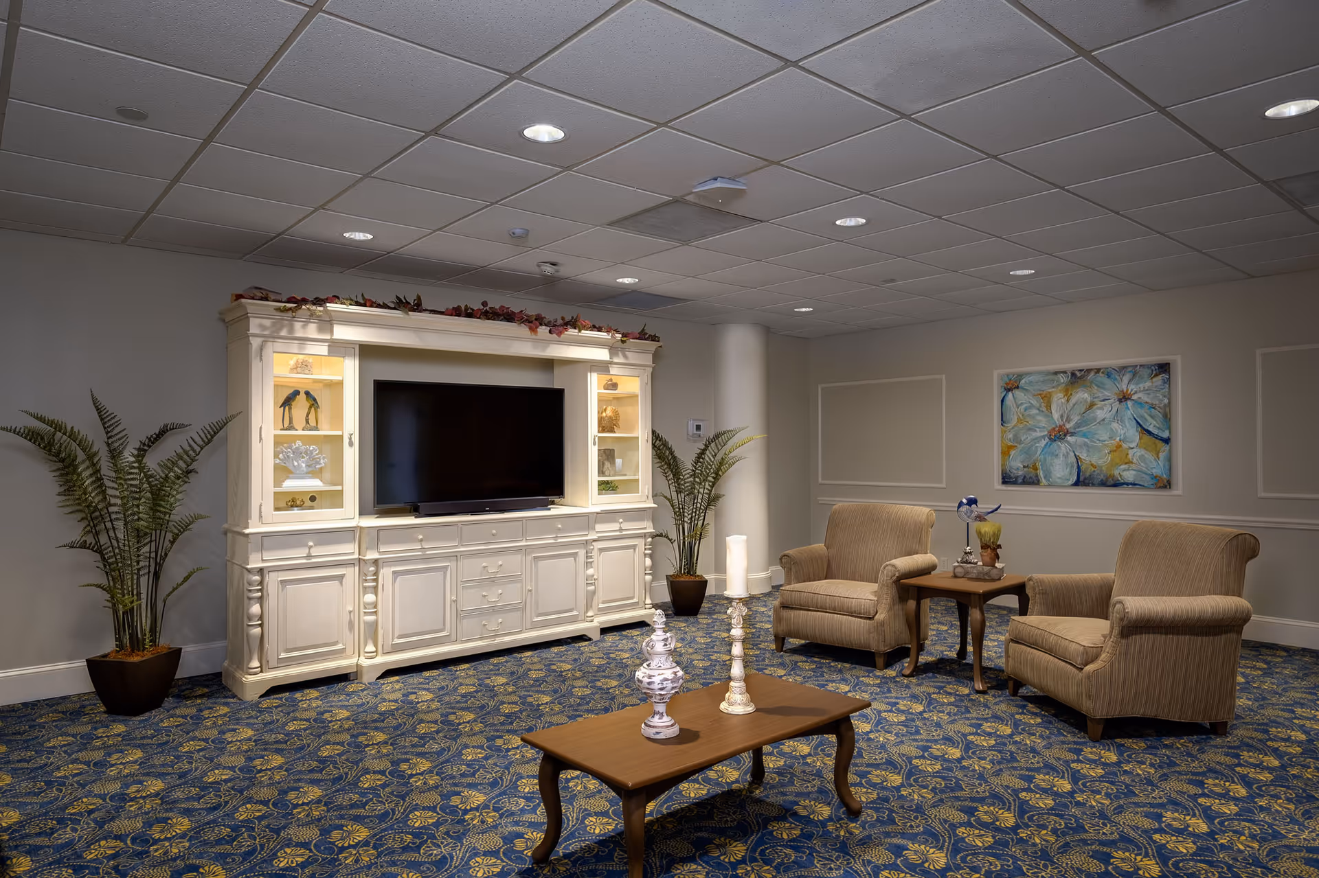 A cozy living room area with two beige upholstered armchairs and a wooden side table between them, decorated with a small plant and a bird figurine. In front of the chairs is a wooden coffee table with two decorative candle holders. Against the wall is a large white entertainment center with a flat-screen TV and decorative items inside glass cabinets. The room has blue and yellow patterned carpet, potted plants on either side of the entertainment center, and a floral painting on the wall.