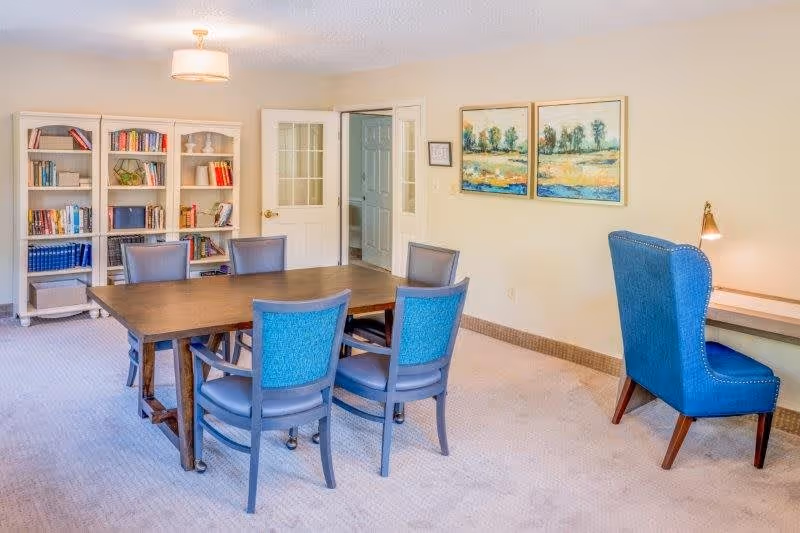 A well-lit room with a wooden table surrounded by six blue cushioned chairs. In the background, there are two white bookshelves filled with books and decorative items. On the right side, there is a blue upholstered chair next to a small desk with a lamp. The walls are beige with two framed landscape paintings hanging above the desk. A door with glass panels is partially open, revealing a hallway.