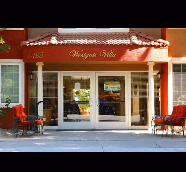 Entrance of Westgate Villa Assisted Living facility with double glass doors, red cushioned chairs on either side, and a red tiled roof above the doorway. The building number 5425 is displayed on the left side of the entrance.
