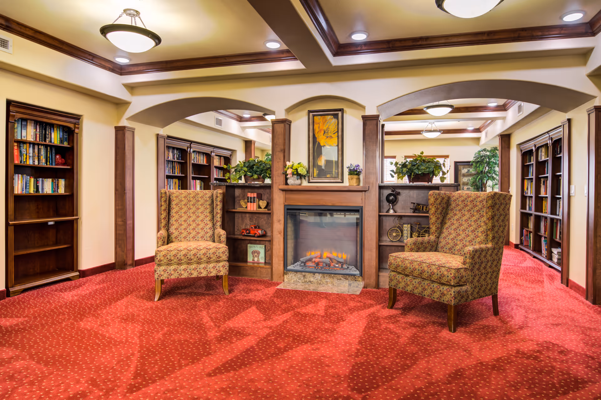 Cozy common room with two patterned armchairs facing a central fireplace flanked by bookshelves and warm lighting.