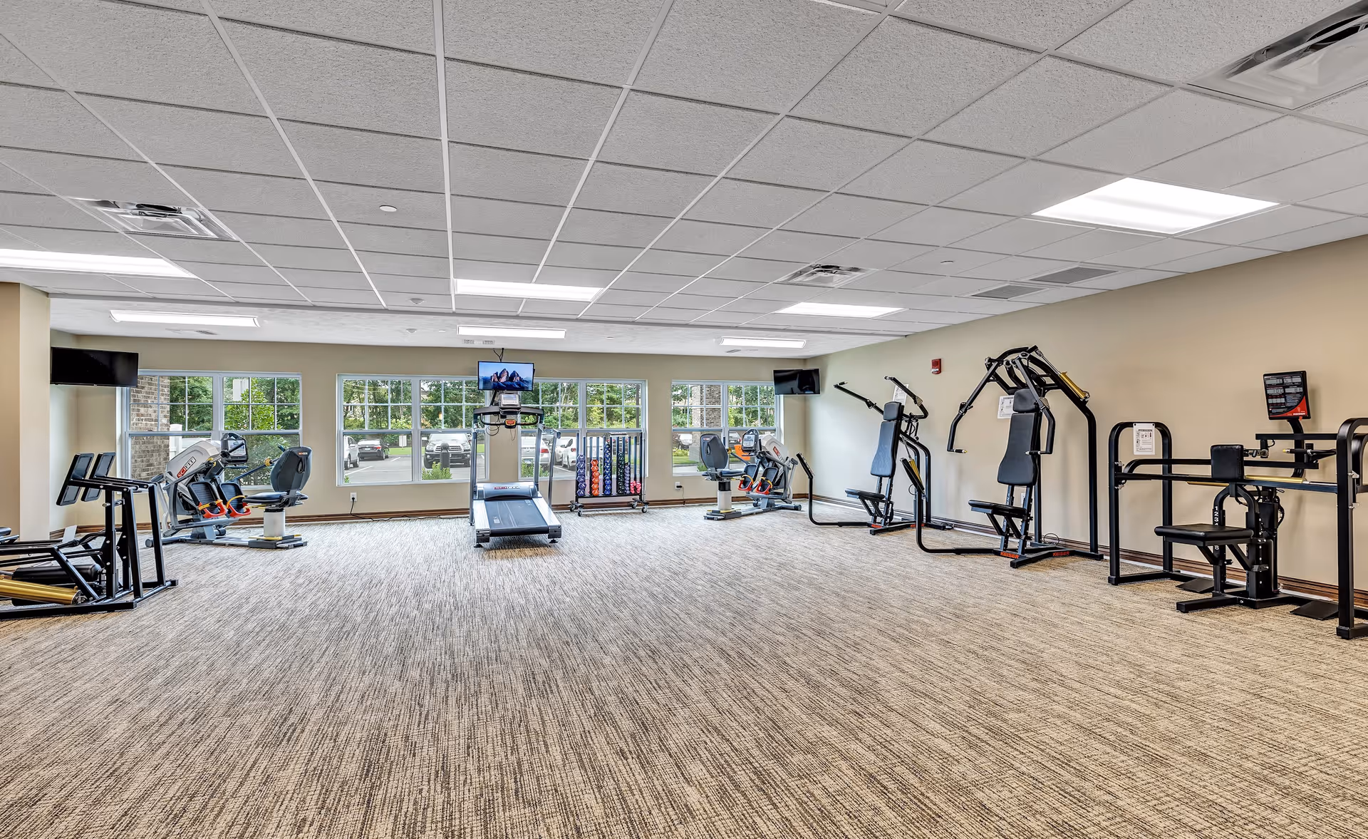 Well-lit exercise room with treadmills, weight machines, free weights, and large windows overlooking the parking lot.