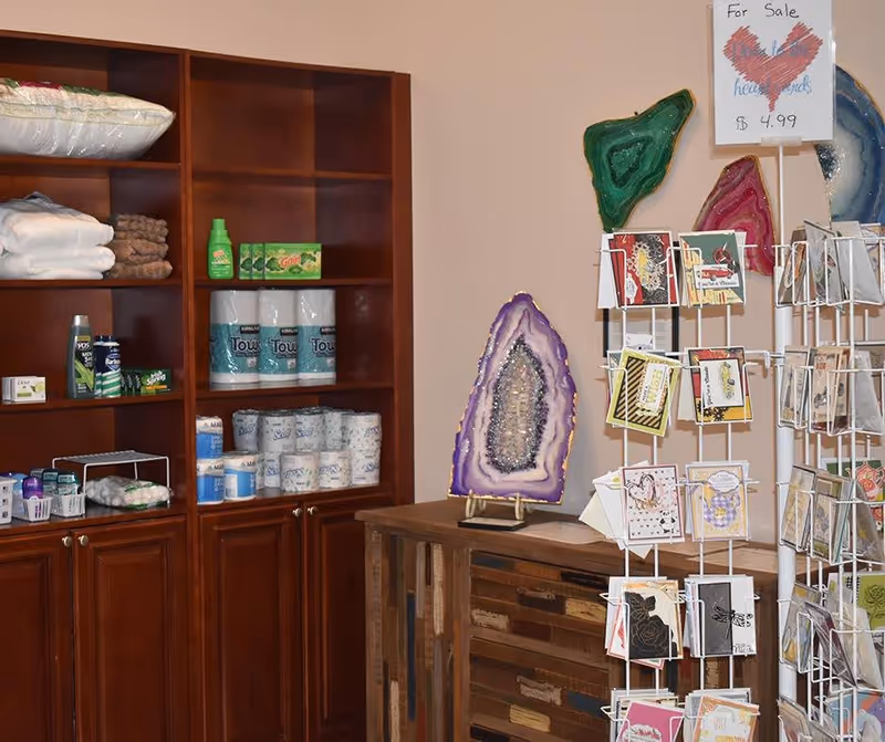 Interior view of a small retail or supply area with wooden shelves stocked with towels, toiletries, and cleaning products. Next to the shelves is a wooden cabinet displaying colorful geode slices and a rotating rack filled with greeting cards. A sign on the rack indicates cards are for sale at $4.99.