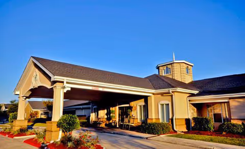 Exterior view of Brookdale Lake Charles senior living facility showing the entrance with a covered driveway, beige building with a small tower structure on the roof, landscaped bushes, and clear blue sky.