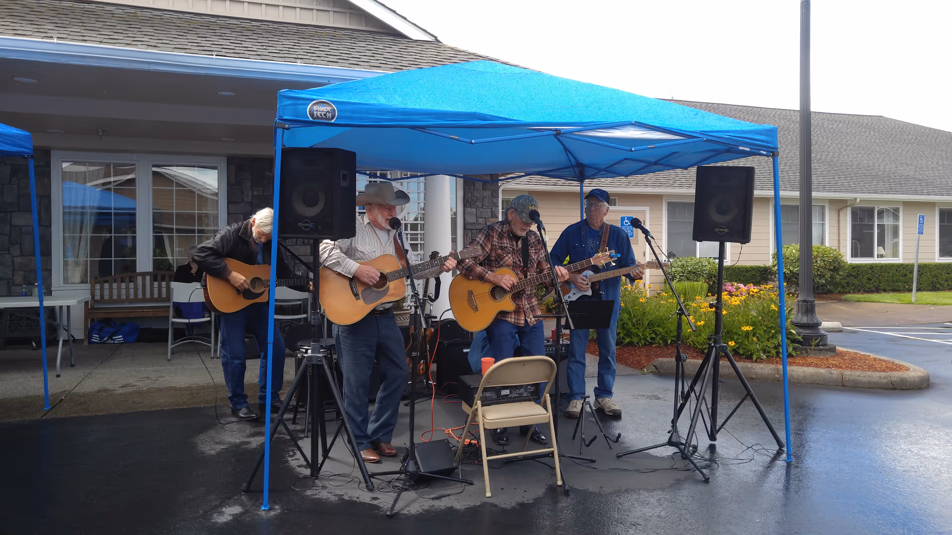 Four men playing guitars and singing under a blue canopy tent outside a building with stone and siding exterior. There are speakers and microphones set up, and a folding chair in front of them. The area appears to be a parking lot with some landscaping and a lamppost nearby.