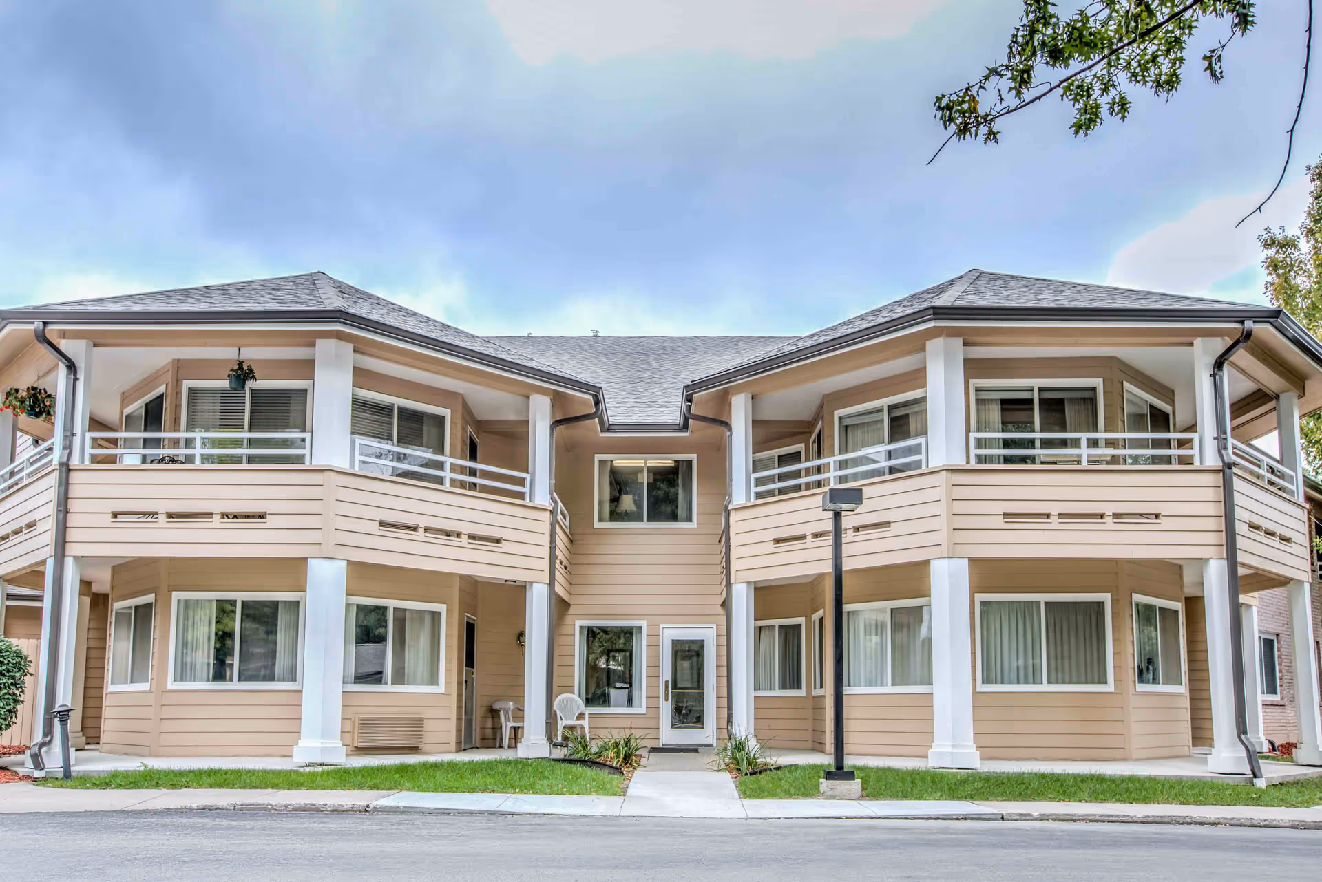 Front exterior view of a two-story beige building with white trim and railings, featuring balconies on the upper floor and large windows on both floors. There is a sidewalk leading to a central entrance door, with green grass and a streetlamp in front.