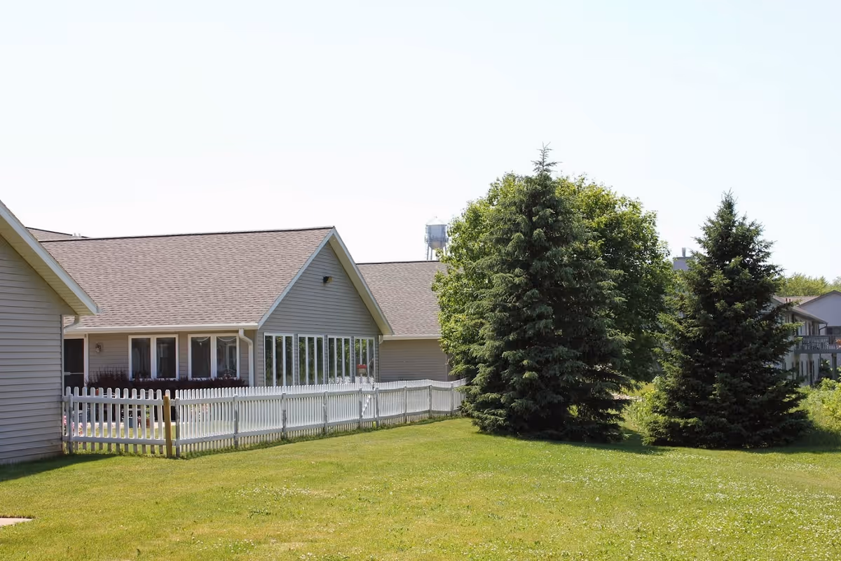 Exterior view of a single-story building with beige siding and a brown shingled roof, surrounded by a white picket fence and green grass. There are two large evergreen trees and a leafy tree in the yard, with a water tower visible in the background under a clear sky.
