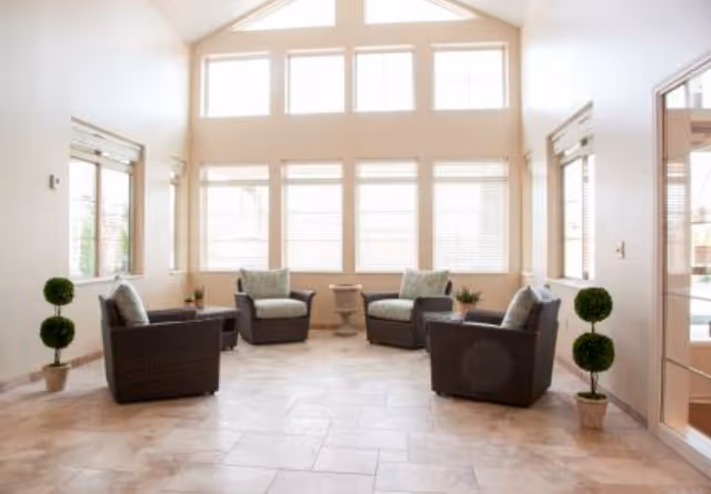 Bright communal lounge with four cushioned wicker chairs arranged around small tables beneath large high windows and potted topiary plants.