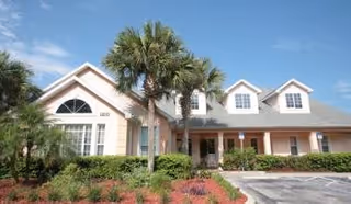 Exterior view of a single-story residential building with a light-colored facade, multiple windows, and a gray shingled roof. There are palm trees and landscaped bushes in front of the building, along with a parking area. The sky is clear and blue.