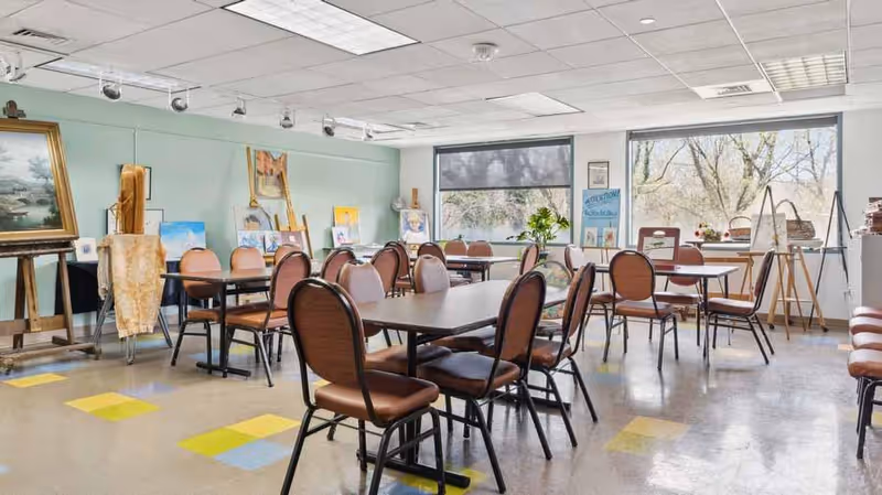 A bright art studio room with multiple tables and chairs arranged for group activities. The walls display various paintings and art supplies, with large windows letting in natural light and showing trees outside.