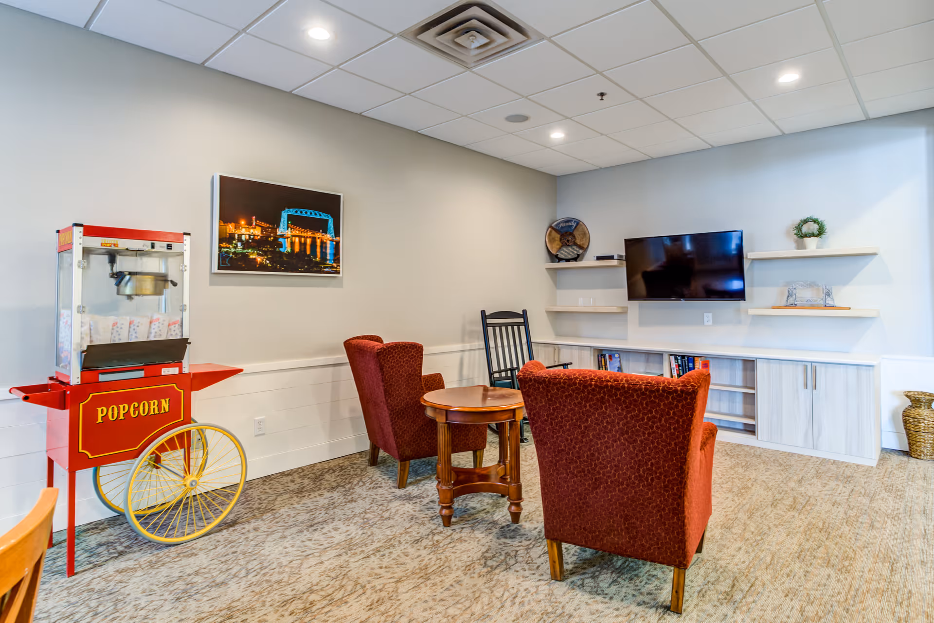 Cozy assisted living lounge with two red upholstered armchairs around a small wooden table, a popcorn cart, wall-mounted TV and shelving.