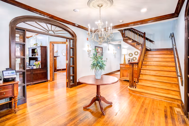 Bright interior space with polished wooden floors and a wooden staircase leading upstairs. A round wooden table with a potted plant is centered under a decorative chandelier. To the left, there is a wooden archway leading to another room with a dark wooden cabinet and a computer. The walls are light-colored with wooden trim and decorative wall sconces.