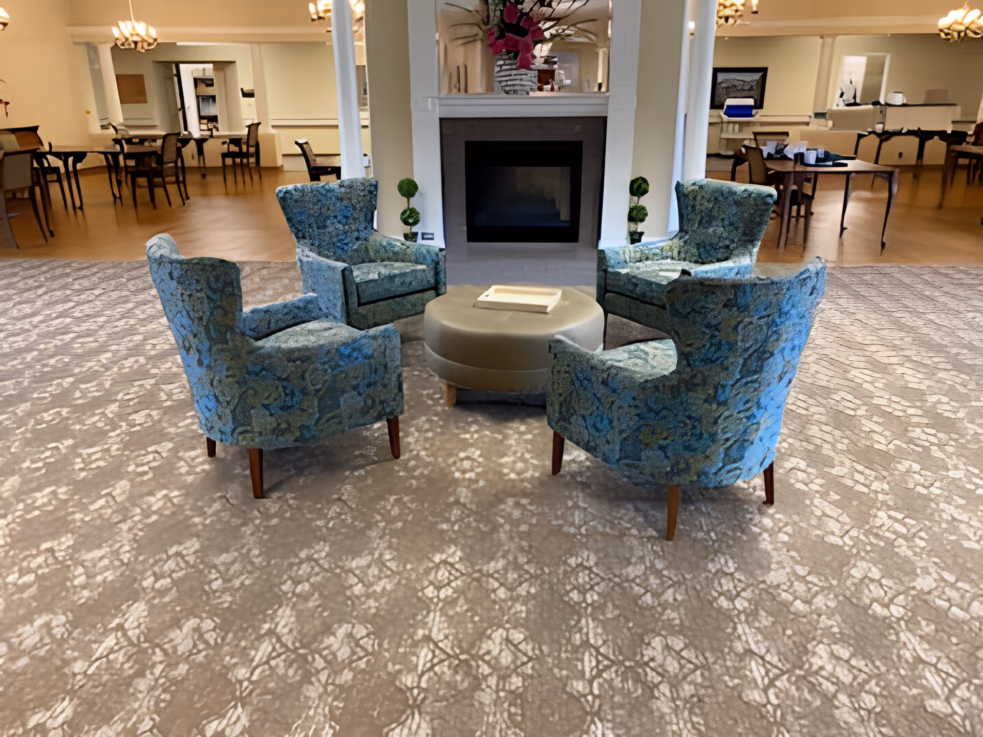 A cozy seating area with four blue patterned armchairs arranged around a round beige ottoman in front of a white fireplace with a floral arrangement on the mantel. In the background, there are multiple dining tables and chairs in a spacious room with beige walls and ceiling lights.