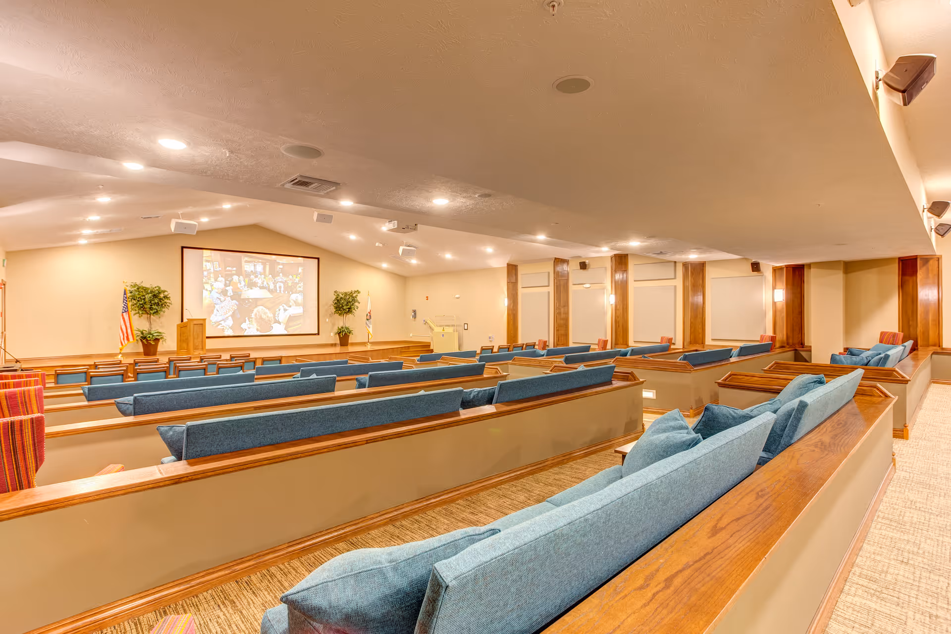 Interior view of a large auditorium or theater room with tiered seating featuring blue cushioned benches and wooden armrests. At the front, there is a stage with a podium, two potted plants, and a large projection screen displaying an image. The room is well-lit with ceiling lights and wall sconces, and has a beige carpeted floor and light-colored walls with wooden panel accents.