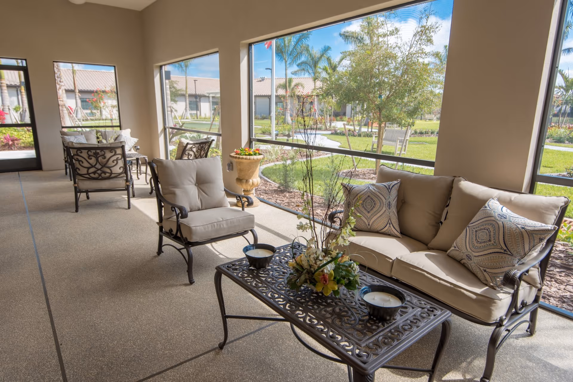 A bright and airy screened-in patio area with cushioned metal chairs and a sofa arranged around a decorative metal coffee table with floral centerpiece and candles. Large windows provide a view of a well-maintained garden with trees, shrubs, and a walking path outside.