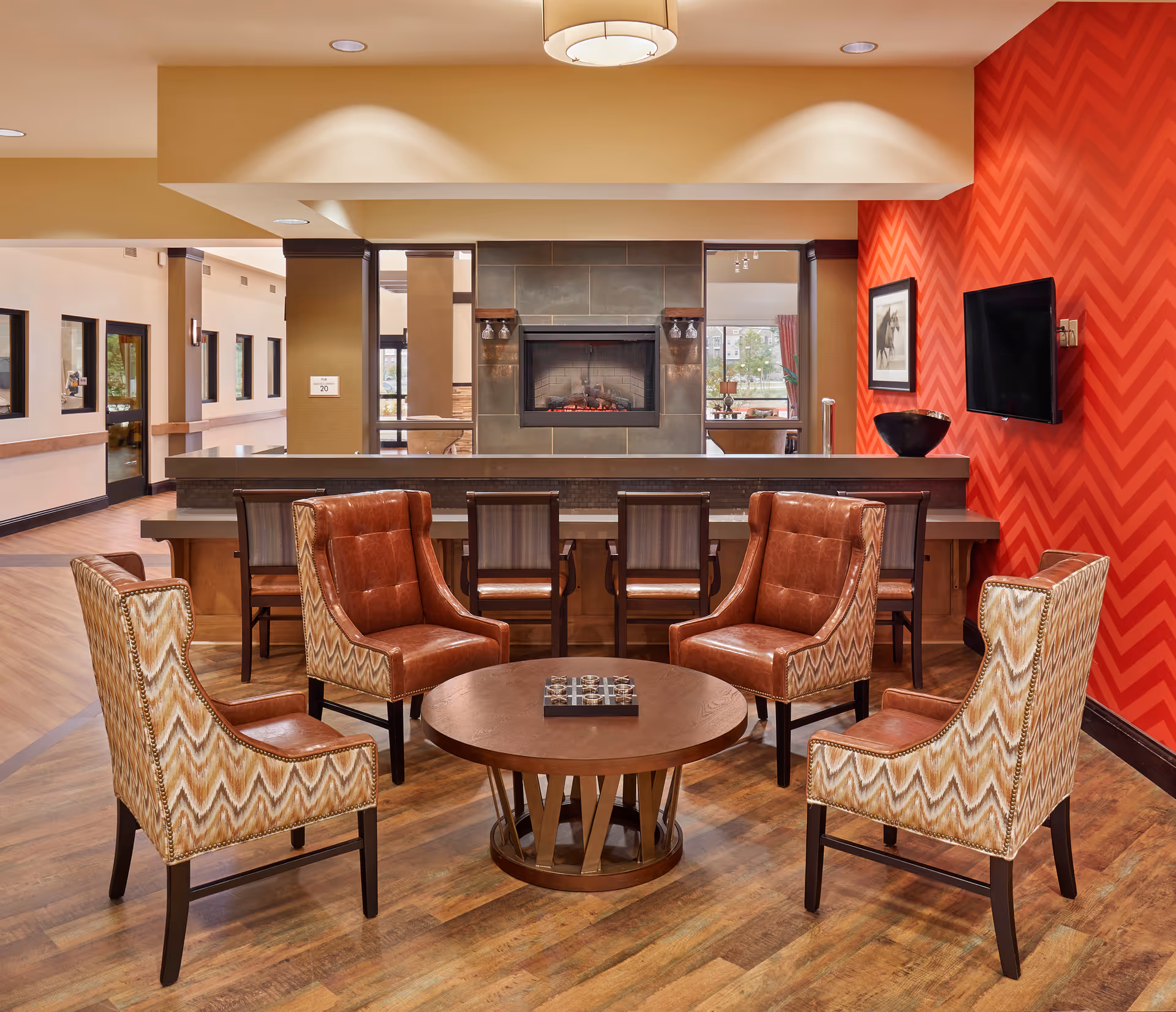 A cozy seating area in a common room featuring four upholstered chairs arranged around a round wooden coffee table. Behind the seating area is a counter with four high chairs and a modern electric fireplace set into a tiled wall. The room has warm wood flooring, a red chevron-patterned accent wall with a mounted flat-screen TV and framed artwork, and large windows letting in natural light.