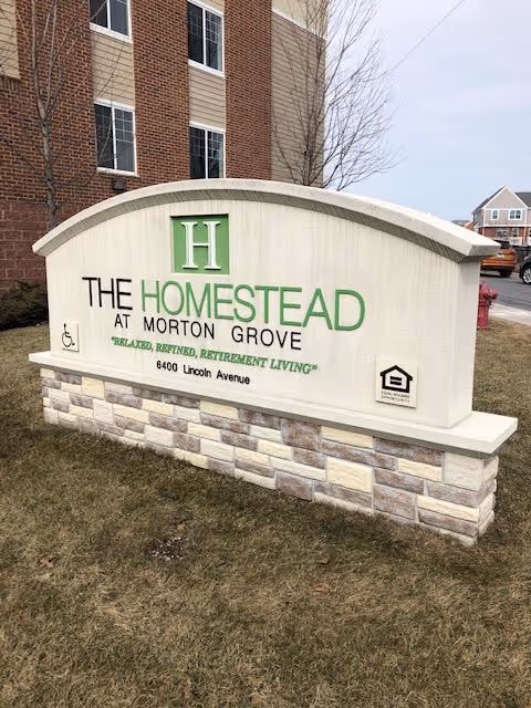 Outdoor sign for The Homestead at Morton Grove, a retirement living facility, located on a grassy area near a brick building and a parking lot.