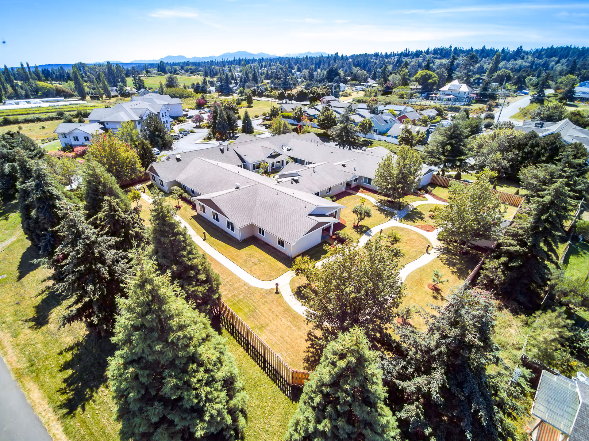 Aerial view of San Juan Villa Memory Care facility surrounded by trees and greenery, showing a single-story building with multiple wings, pathways, and a fenced yard in a suburban neighborhood with other houses and open fields in the background.