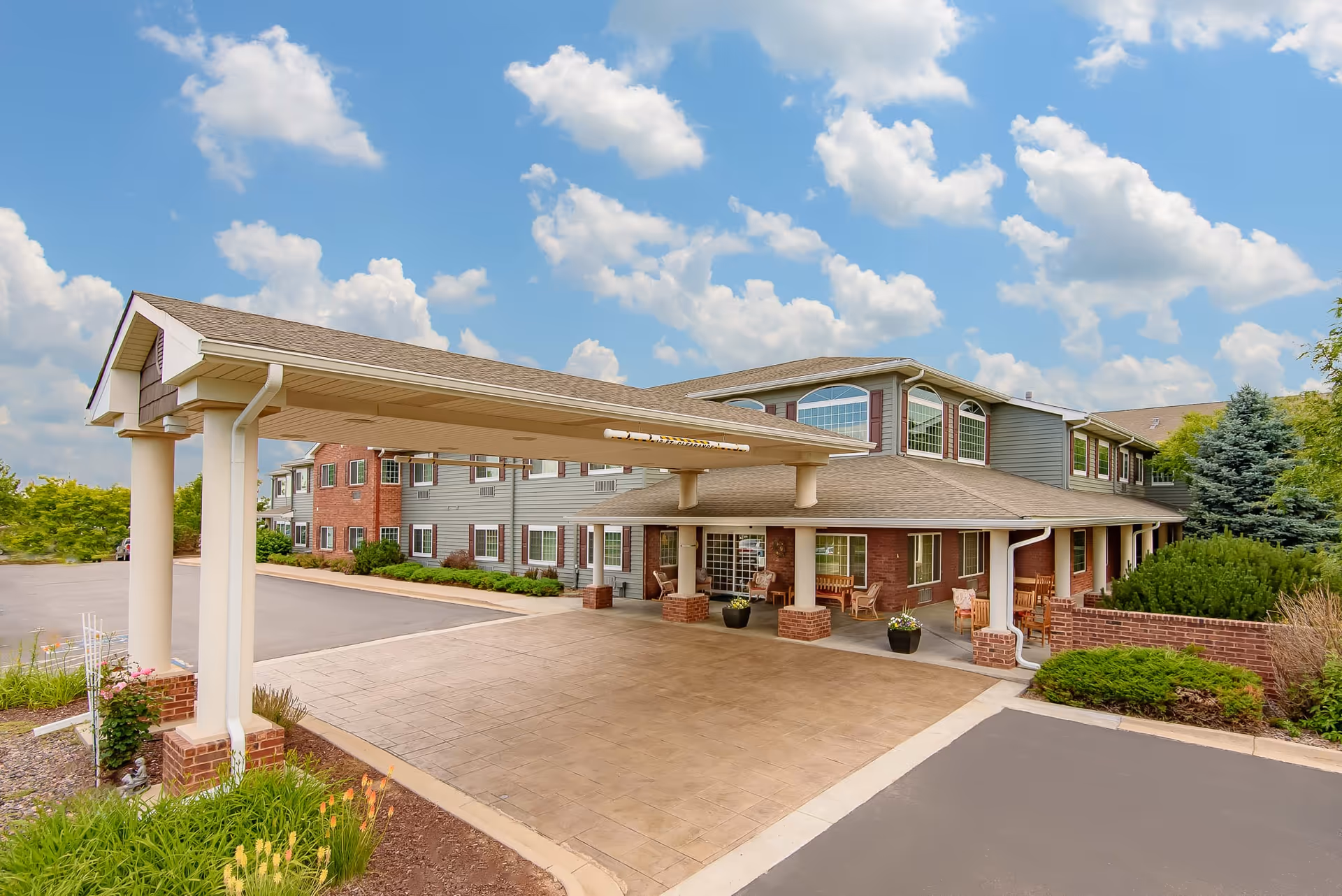 Exterior view of The Bridge at Greeley senior living facility showing a covered entrance with columns, a paved driveway, landscaped greenery, and a two-story building under a partly cloudy sky.