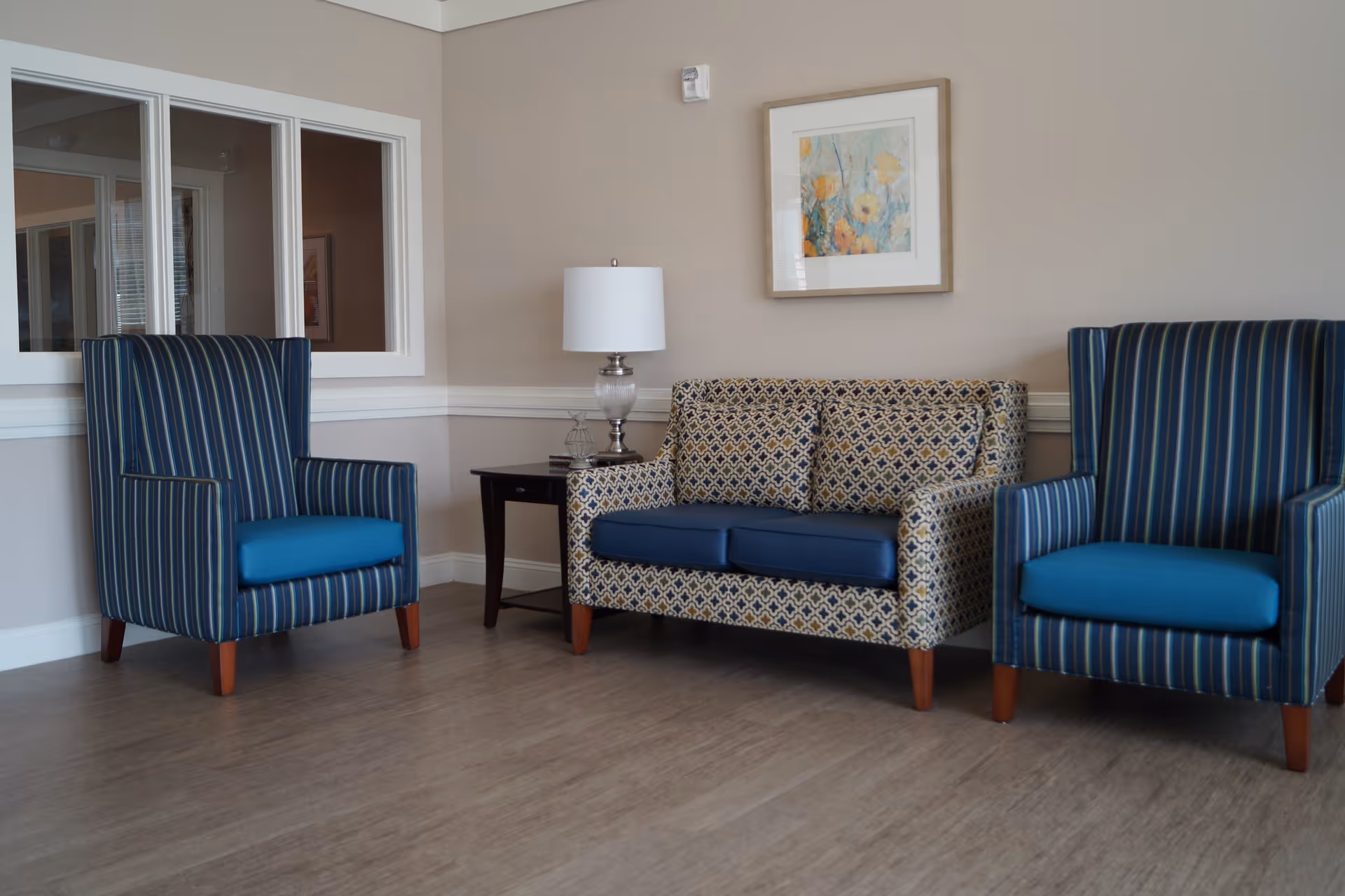 A seating area with two striped armchairs and a patterned loveseat flanking a side table and lamp in a neutral-tone room.