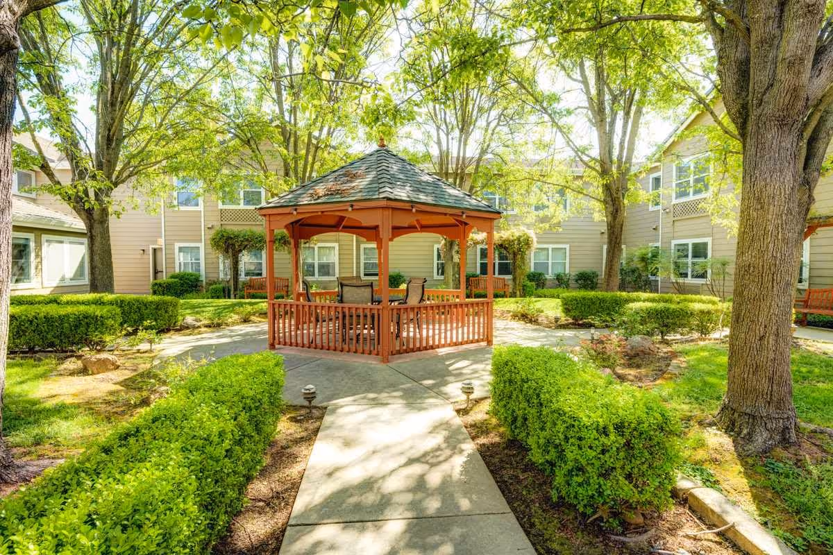 A bright outdoor courtyard area at Vista Roseville Senior Living featuring a wooden gazebo with chairs inside, surrounded by green bushes, trees, and a paved walkway. The senior living facility buildings with multiple windows are visible in the background.