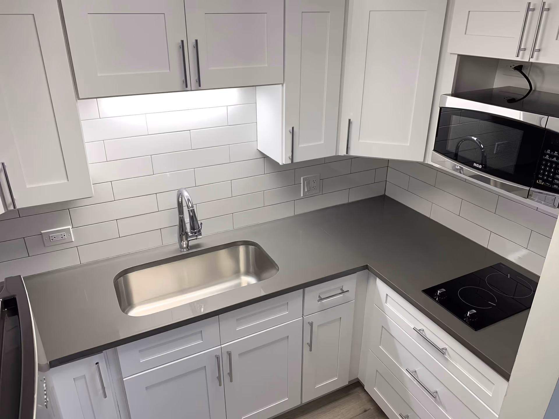 Modern kitchen area with white cabinets, a stainless steel sink, a gray countertop, a built-in microwave, and a black electric stovetop. The backsplash features white subway tiles with gray grout.
