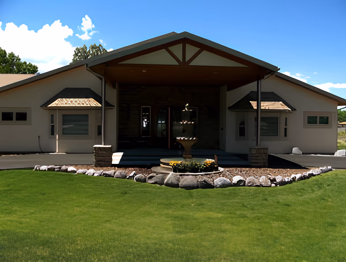 Front exterior view of Grandview Care Lodge Assisted Living building with a covered entrance, stone pillars, a central fountain, and a well-maintained lawn with rocks and flowers in front.