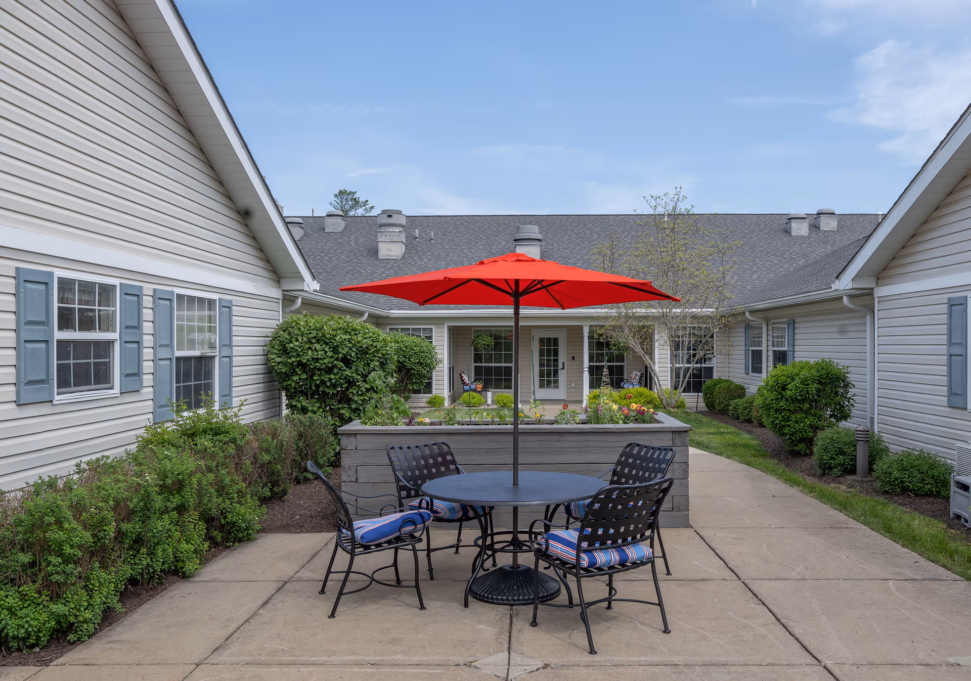 Outdoor courtyard area with a round table and four metal chairs with striped cushions. A large red umbrella is centered over the table. The courtyard is surrounded by beige buildings with blue shutters and green bushes, under a partly cloudy sky.