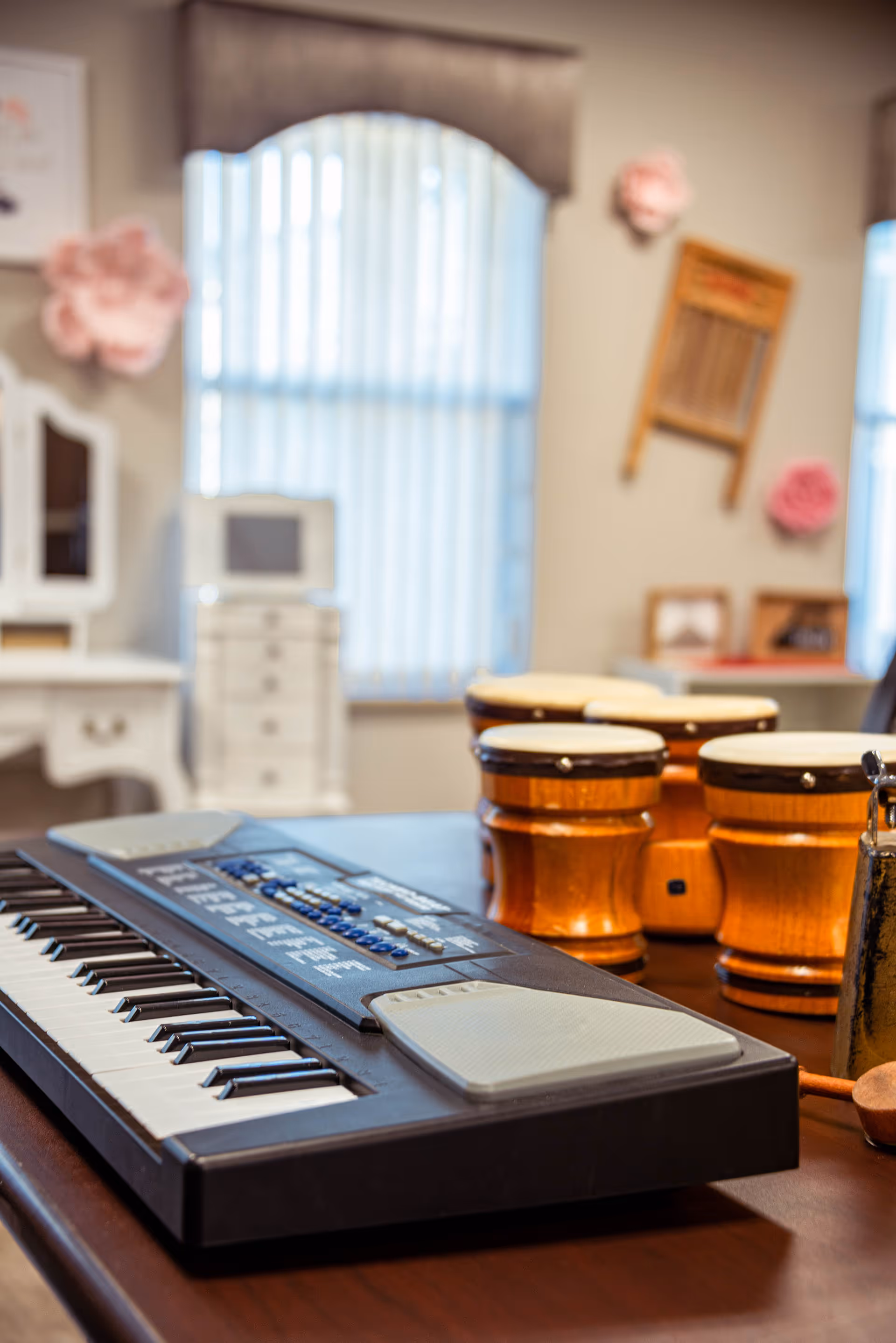 A close-up view of a keyboard and a set of wooden bongo drums on a table in a room with a window covered by vertical blinds. The background includes a white dresser, a mirror, and wall decorations including a washboard and pink flower ornaments.