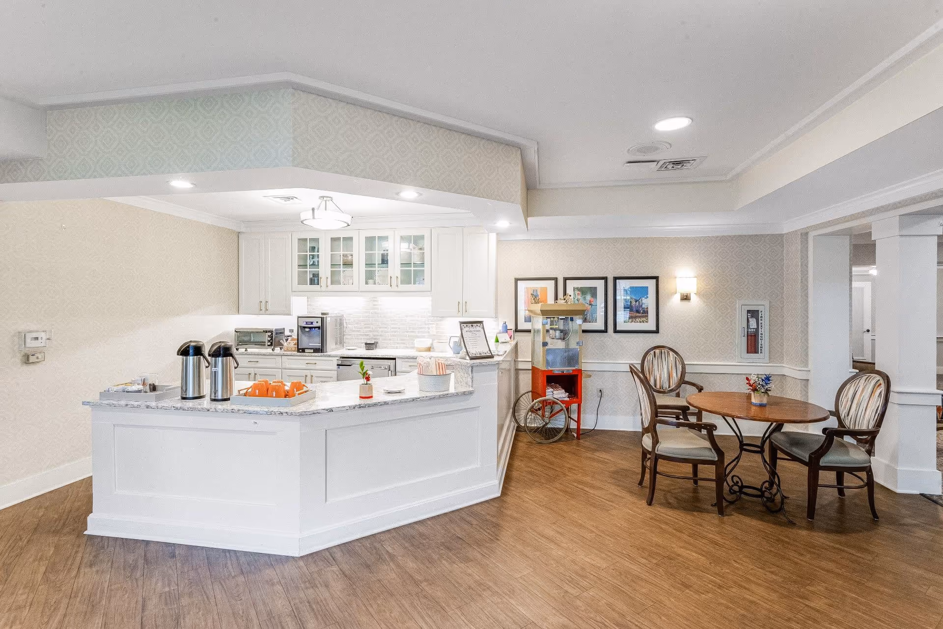 A bright and clean common area with a white kitchen counter featuring coffee dispensers, orange cups, and a small flower vase. Behind the counter are white cabinets with glass doors and a coffee machine. To the right, there is a round wooden table with three cushioned chairs and a popcorn machine on a red stand against the wall. The floor is wooden, and the walls have a light patterned wallpaper with framed pictures and a wall-mounted light.