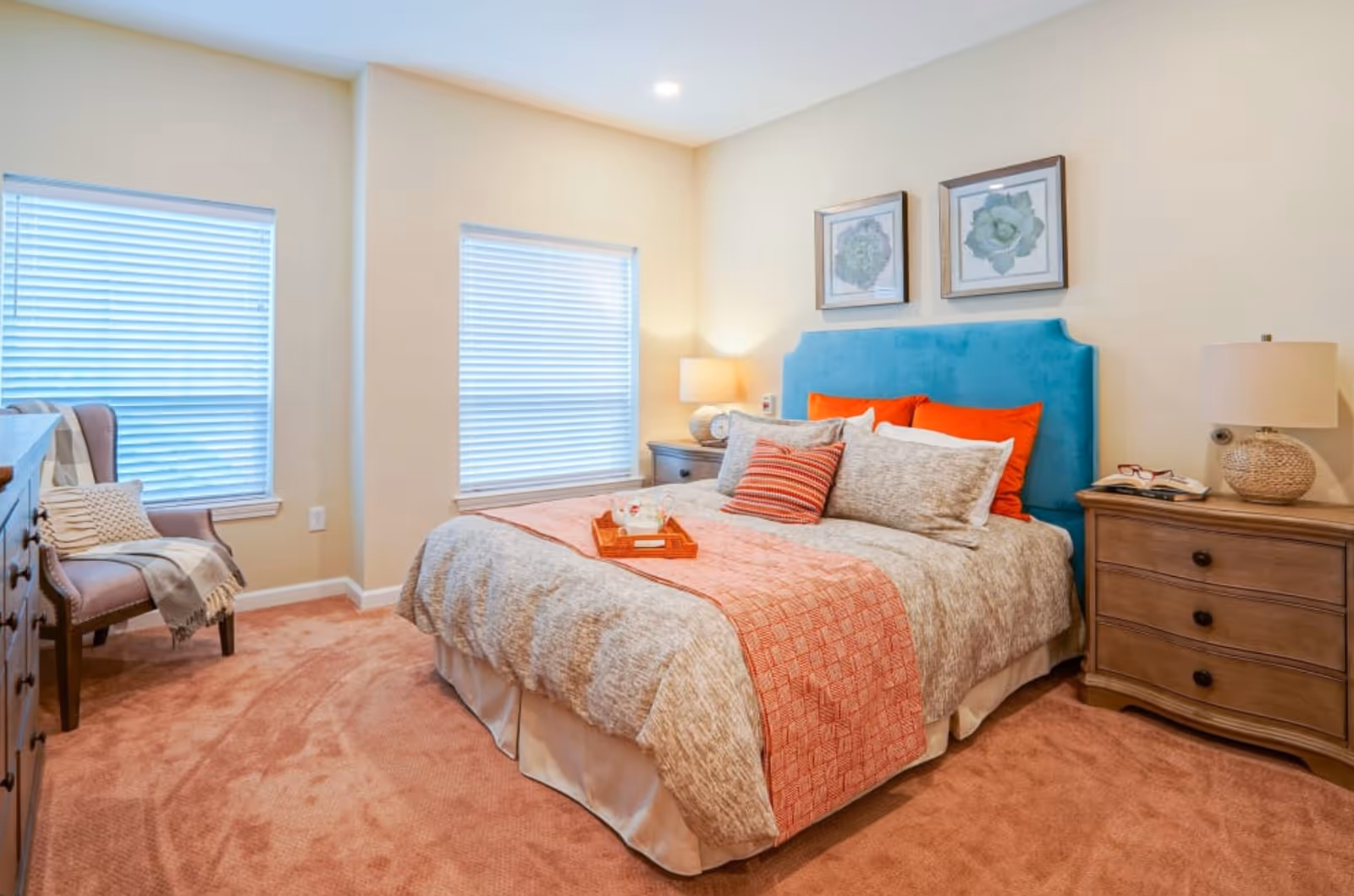 A cozy bedroom with a large bed featuring a blue upholstered headboard, beige and orange bedding, and multiple pillows. There are two framed botanical prints on the wall above the bed. On either side of the bed are wooden nightstands with lamps, one holding a clock and the other a pair of glasses and a book. The room has two windows with white blinds, a pink carpet, and a cushioned chair with a throw blanket in the corner.