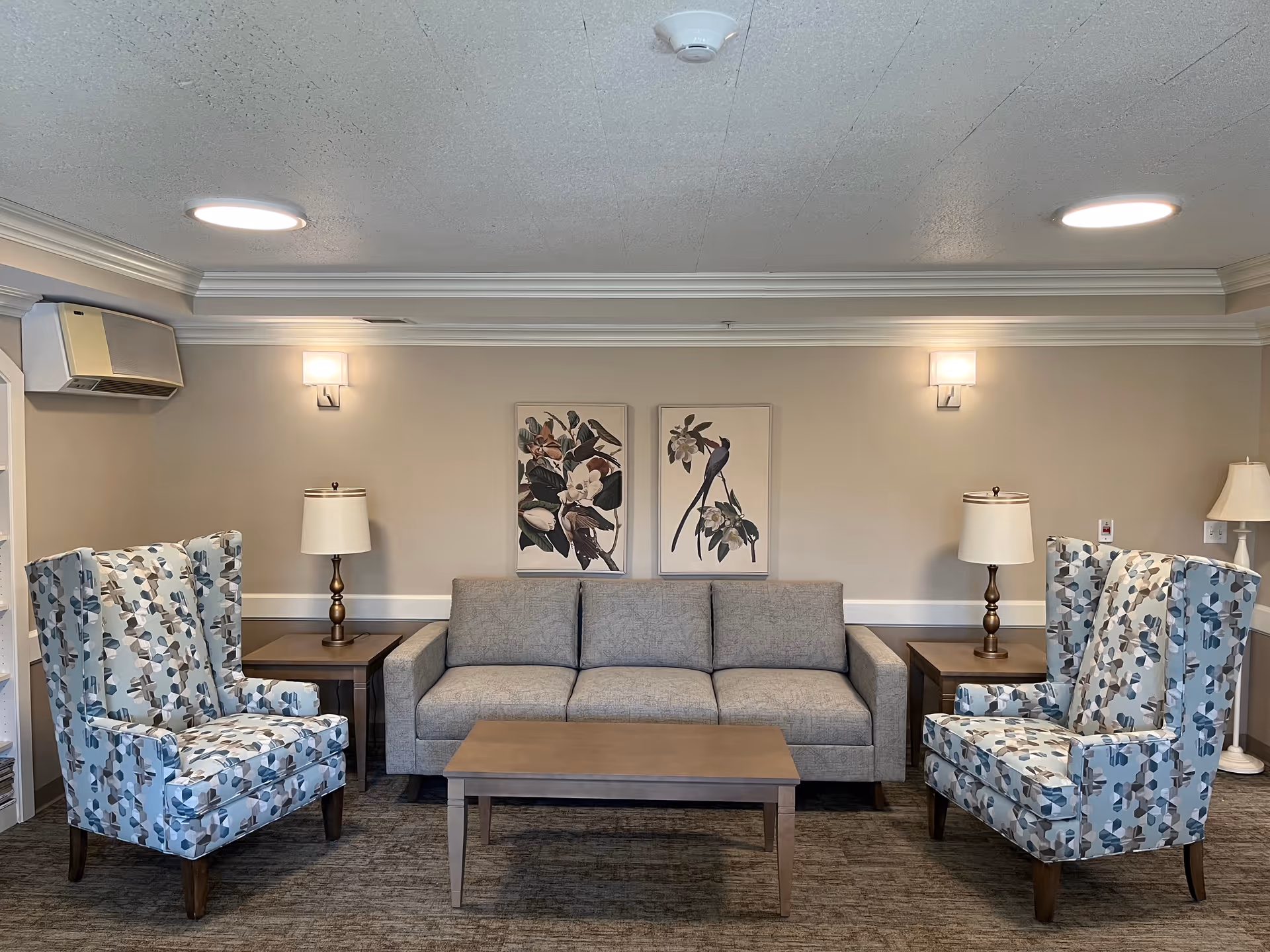 A cozy lounge with a gray sofa centered between two patterned wingback chairs, a coffee table, side tables, lamps, and bird artwork on the wall.