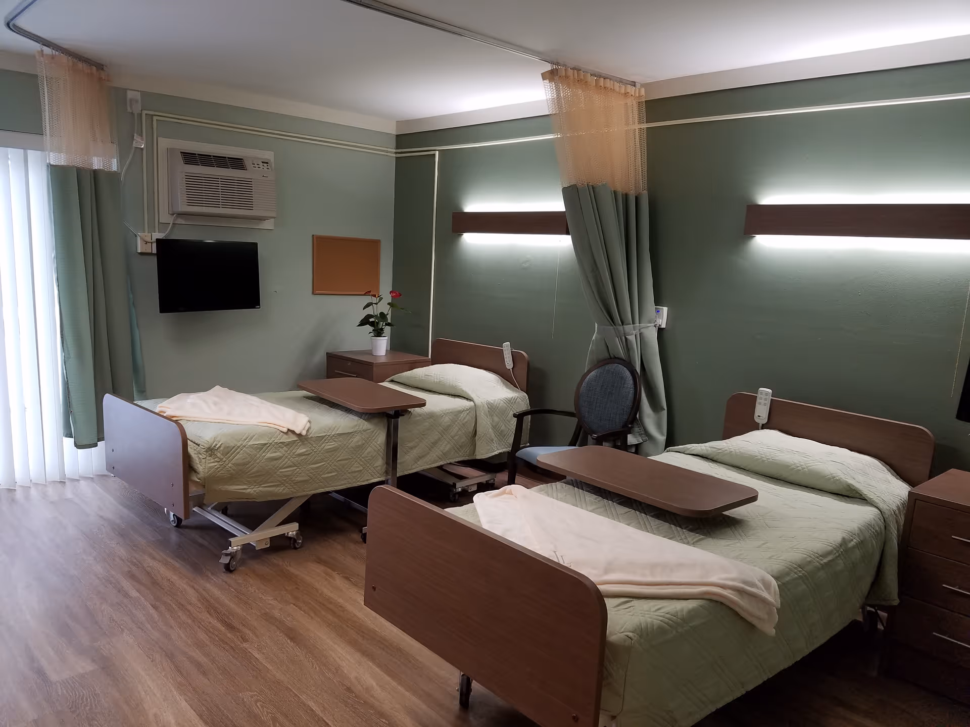 A healthcare facility room with two hospital beds, each with a green quilted blanket and a beige blanket folded at the foot. Each bed has an overbed table and a wall-mounted light above the headboard. There is a chair between the beds, a small nightstand with a potted plant, a wall-mounted TV, and an air conditioning unit. The room has green walls, wood flooring, and a curtain divider between the beds.
