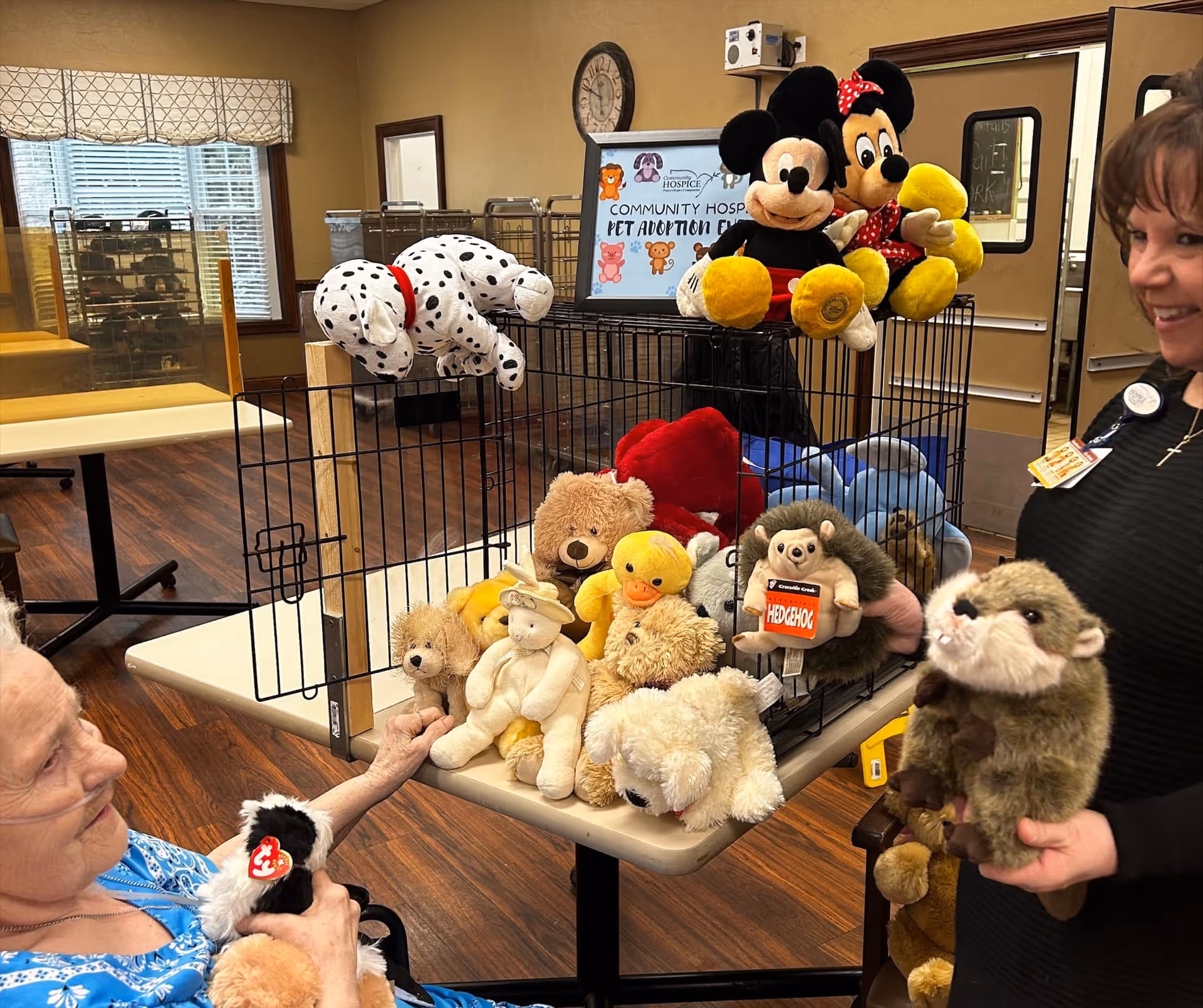 An elderly woman and a staff member interacting with a table full of stuffed animals inside a room with wooden floors and beige walls. The table holds a black wire cage filled with various plush toys including Mickey and Minnie Mouse, a Dalmatian dog, a duck, and other animals. A sign behind the cage reads 'Community Hospice Pet Adoption Event.'