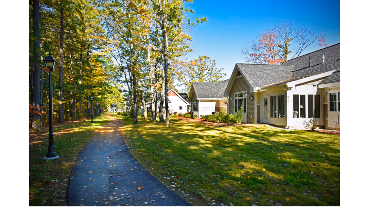 A paved walking path lined with tall trees on the left and residential-style buildings with pitched roofs on the right under a clear blue sky.