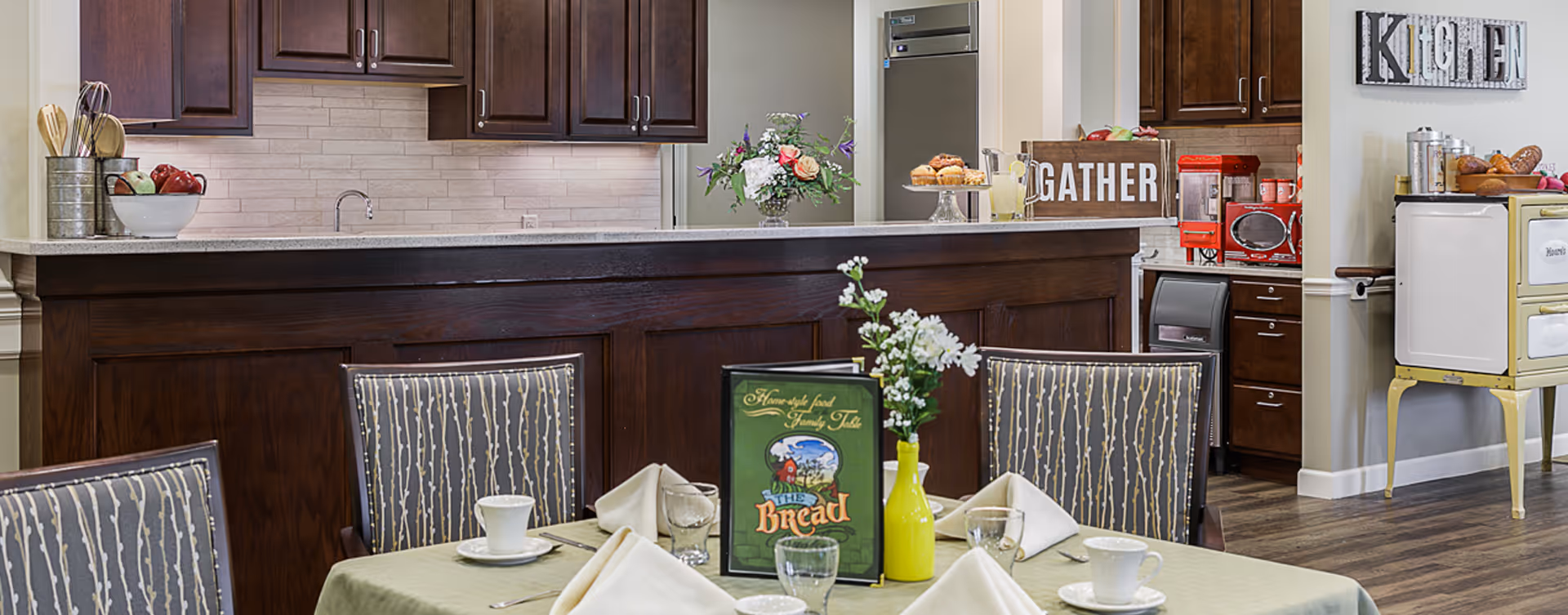 A cozy kitchen and dining area with a table set for a meal, including cups, glasses, napkins, and a menu. The kitchen features dark wood cabinets, a countertop with decorative items like flowers, a sign that says 'GATHER', and a vintage stove. The wall has a decorative sign that reads 'Kitchen'.