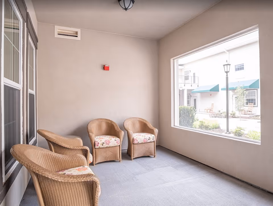 A covered patio area with four wicker chairs that have floral cushions. The patio has beige walls and a large open window looking out to an outdoor courtyard with green awnings and a lamppost.