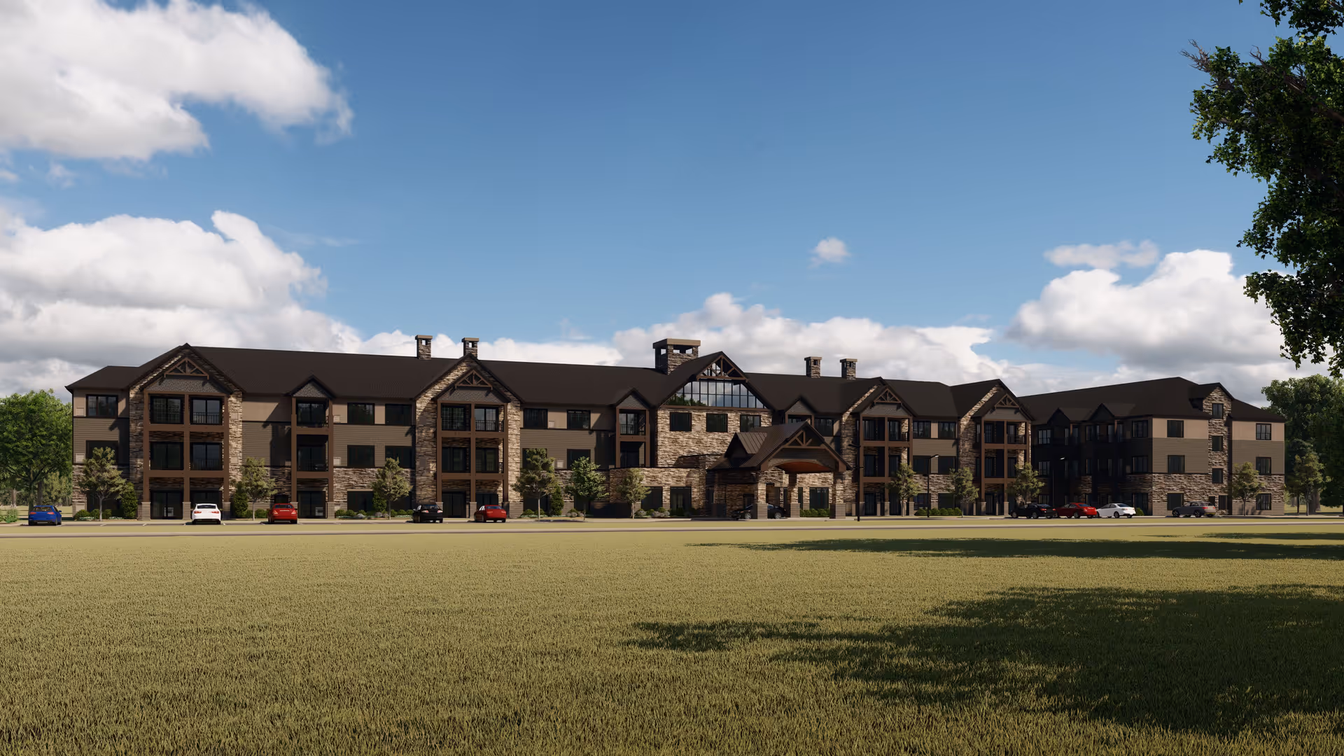 Wide exterior view of a large three-story senior living facility building with stone and wood accents, multiple windows, and a covered entrance. Several cars are parked in front of the building under a partly cloudy blue sky.