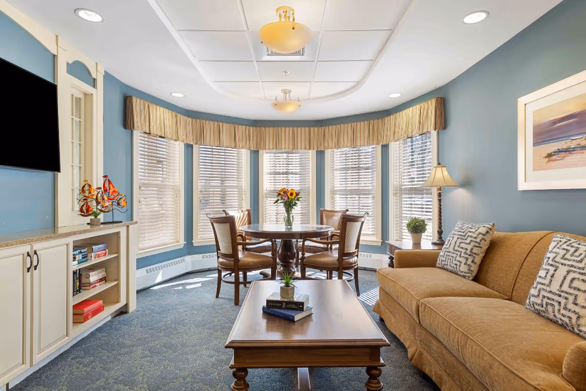A cozy living room with blue walls and a large bay window with blinds and valances. The room features a tan sofa with patterned pillows, a wooden coffee table with books and a small plant, a round wooden dining table with four chairs, a side table with a lamp and plant, a wall-mounted flat screen TV, and a built-in cabinet with shelves holding books and decorative items.