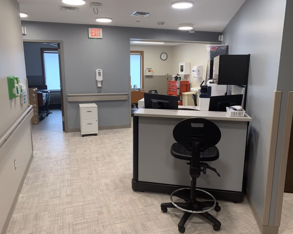 A clean nurses' station and hallway inside a senior care facility with a desk, computer monitors, a stool, hand sanitizer dispenser, and an exit sign.