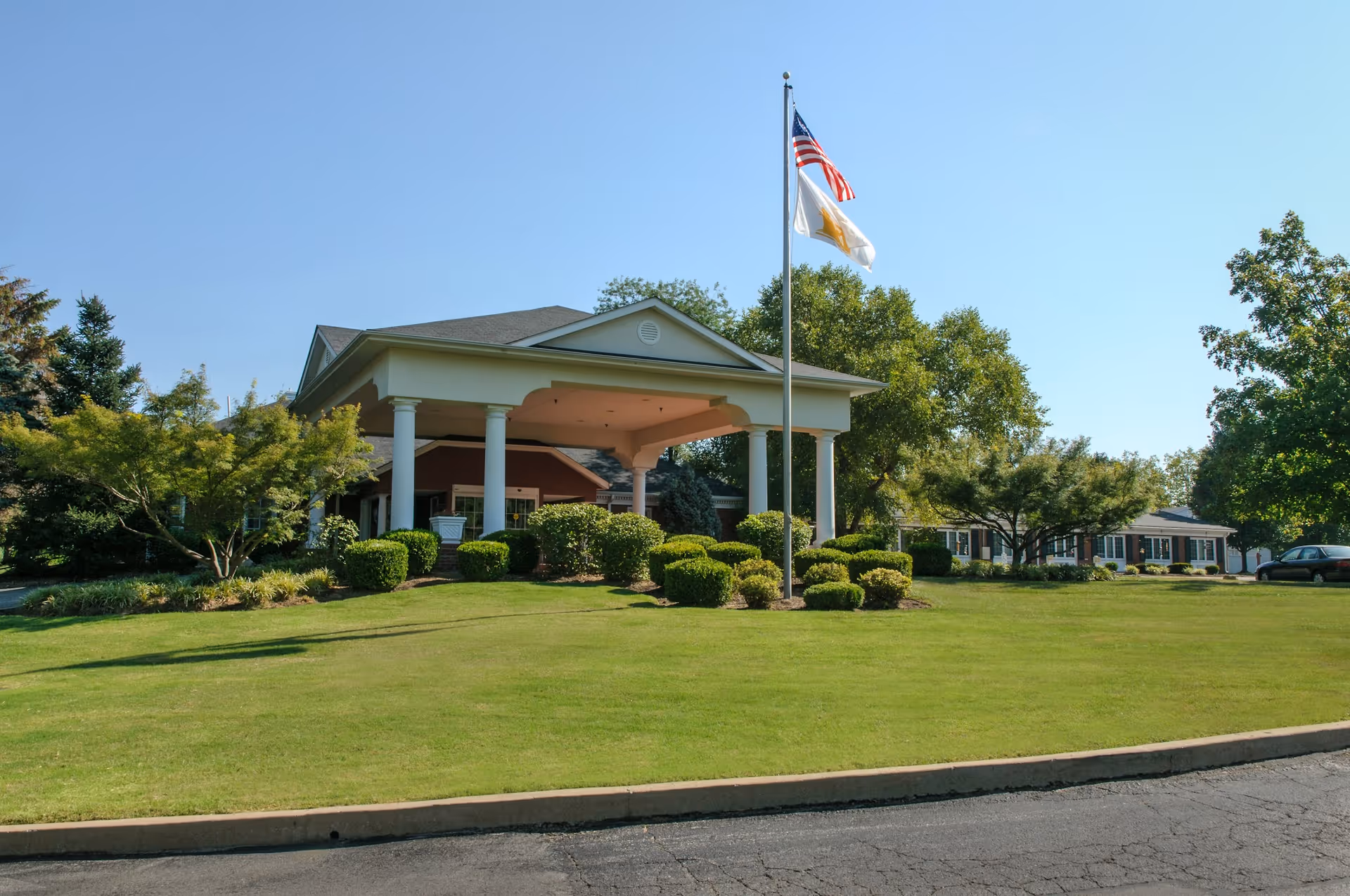 Front entrance of a senior living facility with a covered porte-cochère, flagpoles, and a well-maintained lawn and shrubs.